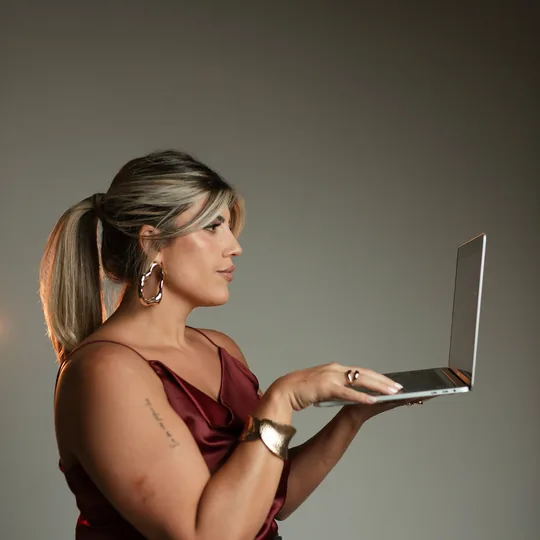 Anndrêa Franco de perfil, blusa bordô, segurando um notebook aberto em estúdio com iluminação lateral