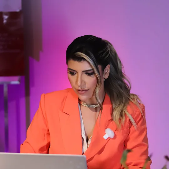 Anndrêa Franco de blazer coral trabalhando no notebook durante evento, com fundo iluminado em roxo