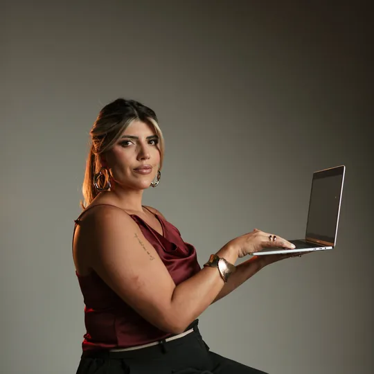 Anndrêa Franco sentada em banqueta, blusa bordô, com notebook nas mãos e olhar para a câmera