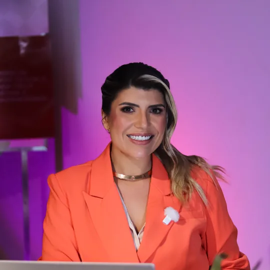 Anndrêa Franco de blazer coral, sorrindo atrás de um notebook durante gravação com fundo iluminado em roxo